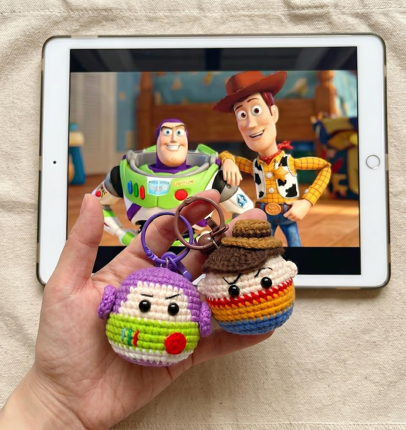 Puede incluir: Dos llaveros de ganchillo hechos a mano con personajes de la pel&iacute;cula de animaci&oacute;n "Toy Story". Un llavero representa a un extraterrestre morado y verde, y el otro a un personaje con un sombrero marr&oacute;n y una camisa a rayas de colores. Los llaveros se sostienen frente a una tableta que muestra una escena de la pel&iacute;cula.