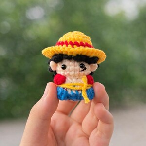 May include: A small, crocheted figure of a character wearing a yellow hat with red trim, a red shirt, blue pants, and a yellow sash. The figure has black hair and is held in a hand against a blurred green background.
