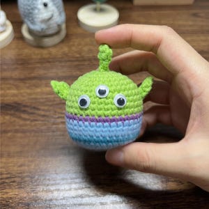 Puede incluir: Un juguete alien&iacute;gena hecho a mano, tejido a ganchillo en hilo verde, azul y morado. El alien&iacute;gena tiene tres ojos y orejas peque&ntilde;as. El juguete est&aacute; sostenido en una mano.