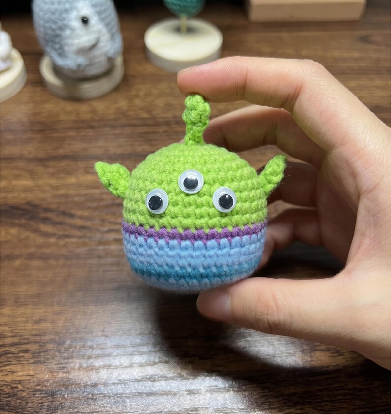 Puede incluir: Un juguete alien&iacute;gena hecho a mano, tejido a ganchillo en hilo verde, azul y morado. El alien&iacute;gena tiene tres ojos y orejas peque&ntilde;as. El juguete est&aacute; sostenido en una mano.