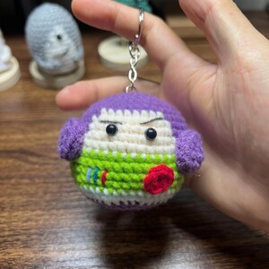 Puede incluir: Un llavero de ganchillo hecho a mano con un personaje redondo con cabeza morada y cara verde. El personaje tiene ojos negros, boca roja y est&aacute; sujeto a un llavero plateado. El llavero est&aacute; sujeto en una mano.