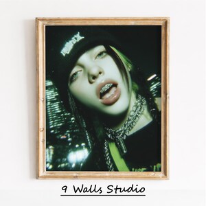 May include: Framed portrait of a person with green-streaked hair, wearing a black beanie with white text, and silver chain necklaces. The person is wearing braces and has a dark background with city lights. The frame is gold.