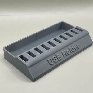 May include: A gray 3D-printed USB holder with a rectangular design. The holder features a series of slots for organizing USB drives and the text "USB Holder" is printed on the side.