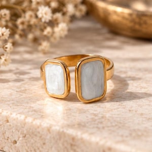 May include: A gold-toned open ring featuring two rectangular settings, each inlaid with a smooth, pale grey enamel stone. The band is polished and metallic, with the two rectangular ends facing each other but not touching, creating an open design.