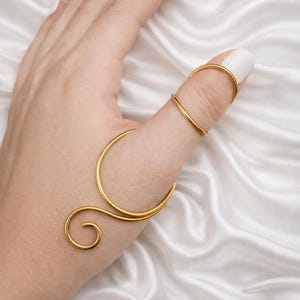 May include: A gold-colored finger ring with a unique design. The ring wraps around the thumb and index finger, featuring a curved band and a spiral detail. The jewelry is set against a white satin background.
