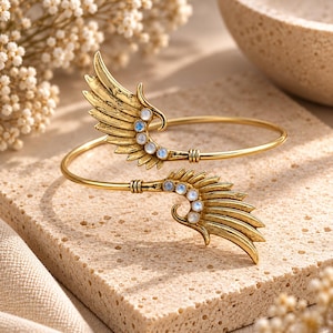 May include: A gold-toned metal cuff bracelet featuring two symmetrical wing-shaped ends. Each wing is detailed with textured feathers and adorned with three small, round, iridescent moonstone-like cabochons set along the inner curve.
