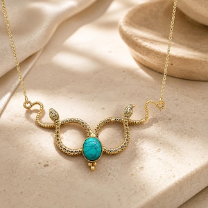 May include: A gold-toned necklace featuring two symmetrical snake figures with textured scales. The snakes meet in the centre to hold an oval-shaped, bright turquoise stone with dark veining. The pendant is attached to a delicate gold-toned chain.
