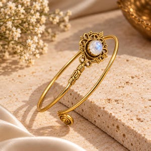 May include: A gold-toned open cuff bracelet featuring a central circular moonstone cabochon set within an ornate, filigree-style metal frame. The bracelet band is smooth and thin, terminating in a small spiral detail at the open end.
