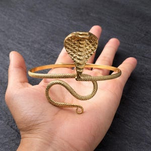 Puede incluir: Un brazalete de serpiente de color dorado. El brazalete presenta una cabeza de cobra y un cuerpo de serpiente enrollado. La cabeza de la cobra está levantada y el cuerpo de la serpiente se enrolla alrededor de la muñeca. El brazalete es de metal brillante.