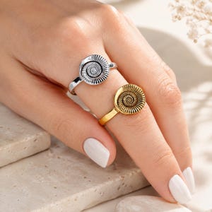 May include: Two rings featuring a circular ammonite fossil design. One ring is finished in a polished silver tone, while the other is finished in a gold tone. Both rings have a detailed spiral pattern radiating from the centre of the circular face.