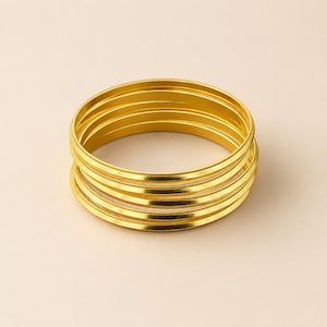 以下が含まれることがあります： 金色の金属製の細い円形バングル5本のスタック。各バングルは、滑らかで光沢のある表面と一貫した丸みを帯びたプロファイルが特徴です。ブレスレットは整然と垂直に積み重ねられており、反射する金属仕上げが際立っています。