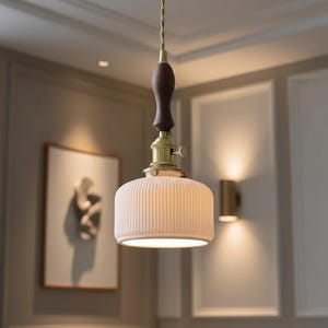 May include: A pendant light fixture with a cream-colored, ribbed ceramic shade. The fixture has a dark brown wooden handle, brass accents, and a twisted cord. The light is hanging in a room with a neutral color palette.