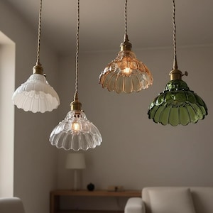 May include: Four pendant lights with scalloped glass shades in white, clear, amber, and green. Each light has a brass-colored fixture and a twisted cord. The lights are hanging from the ceiling in a room with neutral-colored walls.