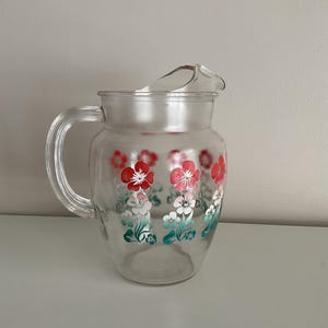 May include: Clear glass pitcher with a handle and spout. The pitcher is decorated with red, white, and teal floral designs. The pitcher is likely used for serving beverages.