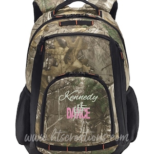 May include: A camouflage backpack with black accents and orange zipper pulls. The front pocket features the name "Kennedy" above the word "DANCE" in pink embroidery. The website address "www.htscreations.com" is printed below.