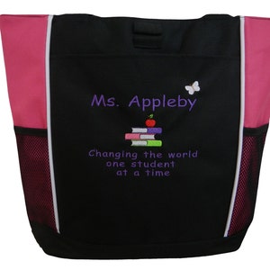 Könnte beinhalten: Schwarze Tragetasche mit rosa Akzenten und einem weißen Reißverschluss. Die Tasche hat eine Netztasche an der Seite und einen gestickten Text mit der Aufschrift "Ms. Appleby Changing the world one student at a time" mit einem Stapel Bücher und einem Apfel darauf und einem Schmetterling.