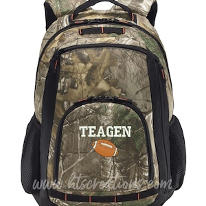 May include: Camouflage backpack with black accents and orange trim. Features the name "TEAGEN" above an orange football graphic. Includes a mesh side pocket and a top handle. The website address "www.htscreations.com" is printed at the bottom.