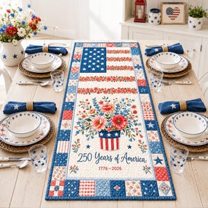 Puede incluir: Un camino de mesa acolchado rectangular con un diseño patriótico de bandera estadounidense, un arreglo floral central en un cubo a rayas y el texto 250 Years of America 1776-2026. El borde consta de cuadrados de patchwork rojos, blancos y azules.