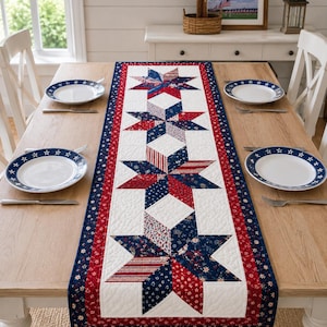 May include: A quilted table runner featuring a repeating star pattern in red, white, and blue fabric. The design includes various patriotic prints, including small floral and striped motifs, bordered by a dark blue fabric with white stars.