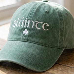 May include: Green baseball cap with the word "sláinte" embroidered in white, along with a small white shamrock. The cap is made of a soft, textured fabric and has a curved brim. The hat is a classic, casual style.