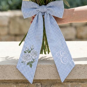 May include: A bouquet of cream and blush roses and white ranunculus, accented with green foliage, is tied with a light blue gingham bow. The bow features embroidered white roses and initials.