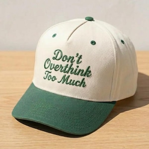 May include: A cream-colored baseball cap with a green brim and embroidered text that reads "Don't Overthink Too Much" in a cursive font. The cap has a green button on top and green eyelets.