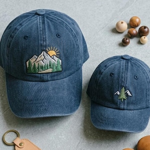 May include: Two blue baseball caps with mountain and tree patches. One cap has a mountain and sun patch, the other has a tree patch. A leather keychain, wooden beads, and brown leather baby shoes are also in the image.