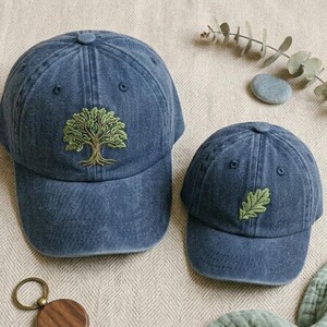 May include: Two blue denim baseball caps. One cap features an embroidered tree design, and the other has an embroidered leaf. The caps are made of a soft, worn denim material. A wooden keychain is in the foreground.
