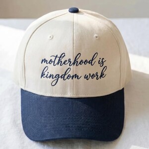 May include: A baseball cap with a cream-colored crown and a navy blue brim. The text "motherhood is kingdom work" is written in navy blue script across the front of the cap. A navy blue button is on top.