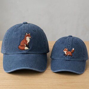 May include: Two blue denim baseball caps featuring embroidered fox designs. The larger cap displays a sitting fox, while the smaller cap features a running fox. Both hats have a structured crown, a curved brim, and a classic six-panel construction.