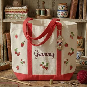 May include: Cream-colored tote bag with red accents and handles, embroidered with strawberries and the word "Grammy." A keychain with a grandmother figure hangs from the handle. The bag is on a wooden surface.