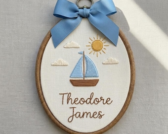 May include: Oval embroidery hoop with a sailboat design. The artwork features a blue sailboat, a yellow sun, and embroidered clouds. The name "Theodore James" is stitched in brown thread. A blue ribbon bow is attached at the top.