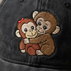 May include: Black baseball cap featuring an embroidered design of two cartoon monkeys hugging. One monkey has a red bow. The monkeys are brown and tan, with black eyes and smiling faces. The cap is made of a dark, possibly denim, material.