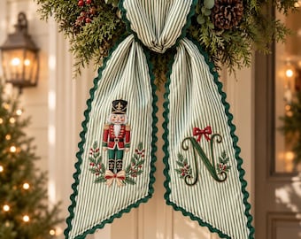 May include: A decorative fabric hanging featuring green and white vertical stripes with a scalloped dark green edge. One side displays an embroidered nutcracker figure, while the other side features a large embroidered letter N with a red bow.