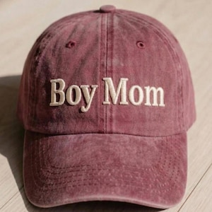 May include: A maroon baseball cap with the embroidered text "Boy Mom" in white. The cap has a curved brim and a fabric strap closure. The hat is made of a soft, washed cotton material.