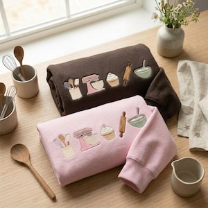 May include: A dark brown sweatshirt and a light pink sweatshirt, each featuring embroidered kitchen motifs including a utensil crock, a stand mixer, a cupcake, a rolling pin, and a mixing bowl. The garments are folded on a wooden surface.