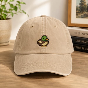 May include: A tan baseball cap featuring a small, embroidered pixel art mallard duck on the front center. The duck design consists of green, brown, yellow, and black thread, creating a blocky, retro-inspired graphic on the solid-colored fabric.