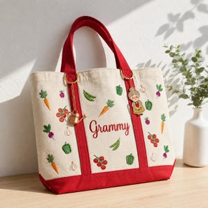 May include: Cream-colored tote bag with red accents and handles, embroidered with vegetables and the word "Grammy." Features a charm of a watering can and a woman. A charming and practical accessory.