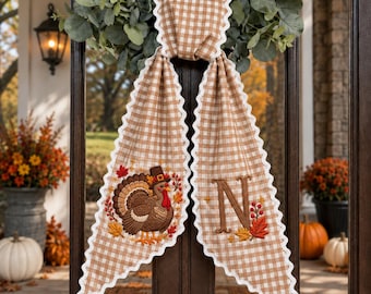 May include: A tan and white gingham fabric door hanger featuring a scalloped white edge. The left side displays an embroidered turkey wearing a pilgrim hat, while the right side features a large embroidered letter N with autumn leaves and berries.