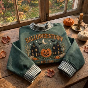 May include: A dark green sweatshirt featuring a gingham collar and cuffs. The front displays embroidered text reading HALLOWEENTOWN EST. 1998, with a central orange jack-o'-lantern, two dark houses, a crescent moon, stars, and bats.