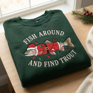 May include: A dark green crewneck sweatshirt featuring an embroidered trout wearing a Santa hat and a belt, wrapped in colorful Christmas lights. The text "FISH AROUND AND FIND TROUT" is embroidered in white above and below the fish graphic.