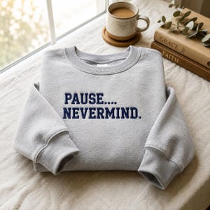 May include: A light gray crewneck sweatshirt featuring the dark blue embroidered text PAUSE.... NEVERMIND. centered on the chest. The garment has long sleeves with ribbed cuffs and a ribbed neckline, showcasing a clean and simple design.