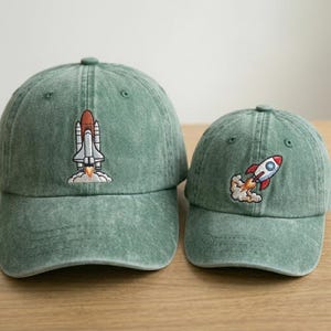 May include: Two sage green baseball caps with a distressed finish. The larger cap features an embroidered white and brown space shuttle launching. The smaller cap features an embroidered red, white, and blue rocket ship launching with orange flames.