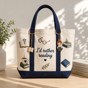 May include: Cream-colored tote bag with navy blue accents and handles. The bag features embroidered images of books, a coffee cup, and the text "I'd rather reading." Gold-colored charms of books and a person reading hang from the handles.