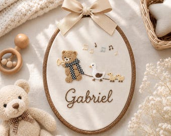 May include: An oval wooden embroidery hoop containing cream-colored fabric with a hand-stitched design of a brown teddy bear playing a trumpet, followed by a white duck and two ducklings. The name Gabriel is embroidered in brown script below.