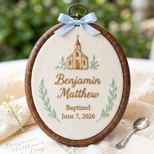 May include: An oval embroidery hoop with a dark wood grain frame, featuring a light blue ribbon bow at the top. The cream fabric displays an embroidered gold church, green leaf sprigs, and the text Benjamin Matthew Baptized June 7, 2026.