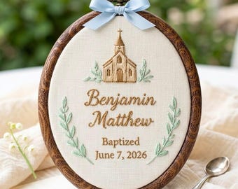 May include: An oval embroidery hoop with a dark wood grain frame, featuring a light blue ribbon bow at the top. The cream fabric displays an embroidered gold church, green leaf sprigs, and the text Benjamin Matthew Baptized June 7, 2026.