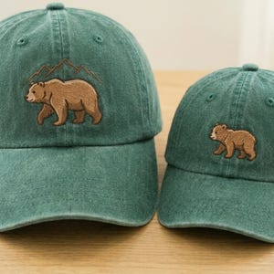 May include: Two forest green baseball caps with a distressed finish. The larger cap features an embroidered brown bear walking beneath mountain outlines. The smaller cap features a single embroidered brown bear centered on the front panel.