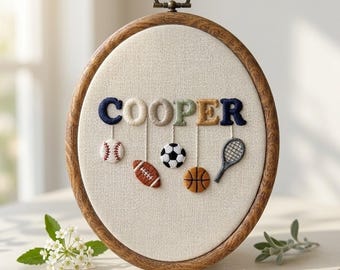 May include: An oval wooden embroidery hoop containing cream fabric with the name COOPER stitched in multi-colored letters. Below each letter hangs a small embroidered sports icon including a baseball, football, soccer ball, basketball, and tennis racket.