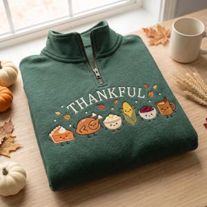 May include: A dark green quarter-zip sweatshirt featuring the word THANKFUL embroidered in white above a row of cute, cartoon-style Thanksgiving food items including a slice of pie, a roasted turkey, mashed potatoes, corn, and a mug.